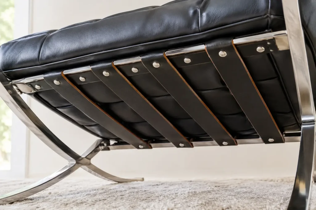 Underside of a black leather Barcelona chair replica showing leather support straps riveted to the stainless steel frame